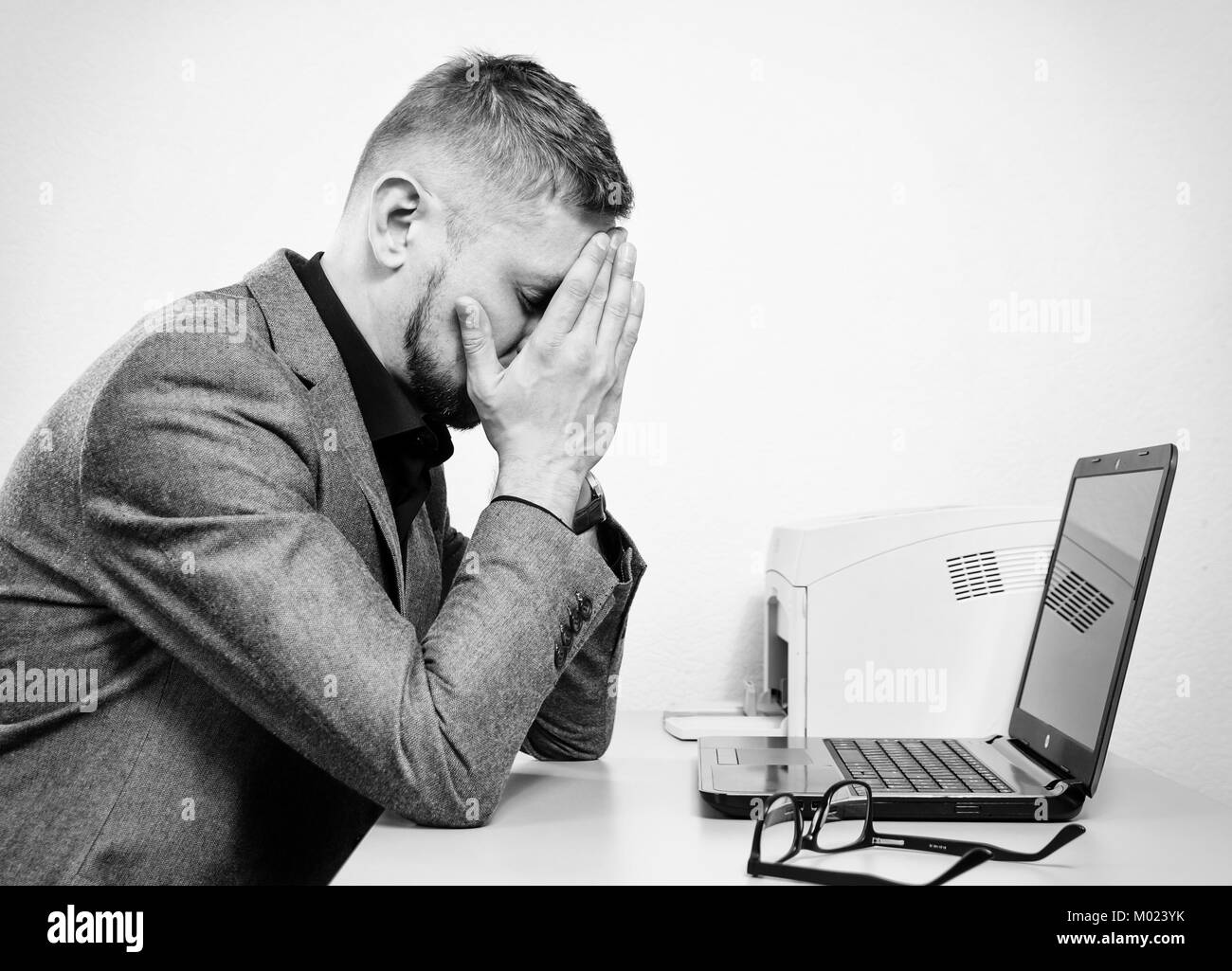 a tired man with his hands on the his face at the table with a laptop in the office. black and white. Stock Photo