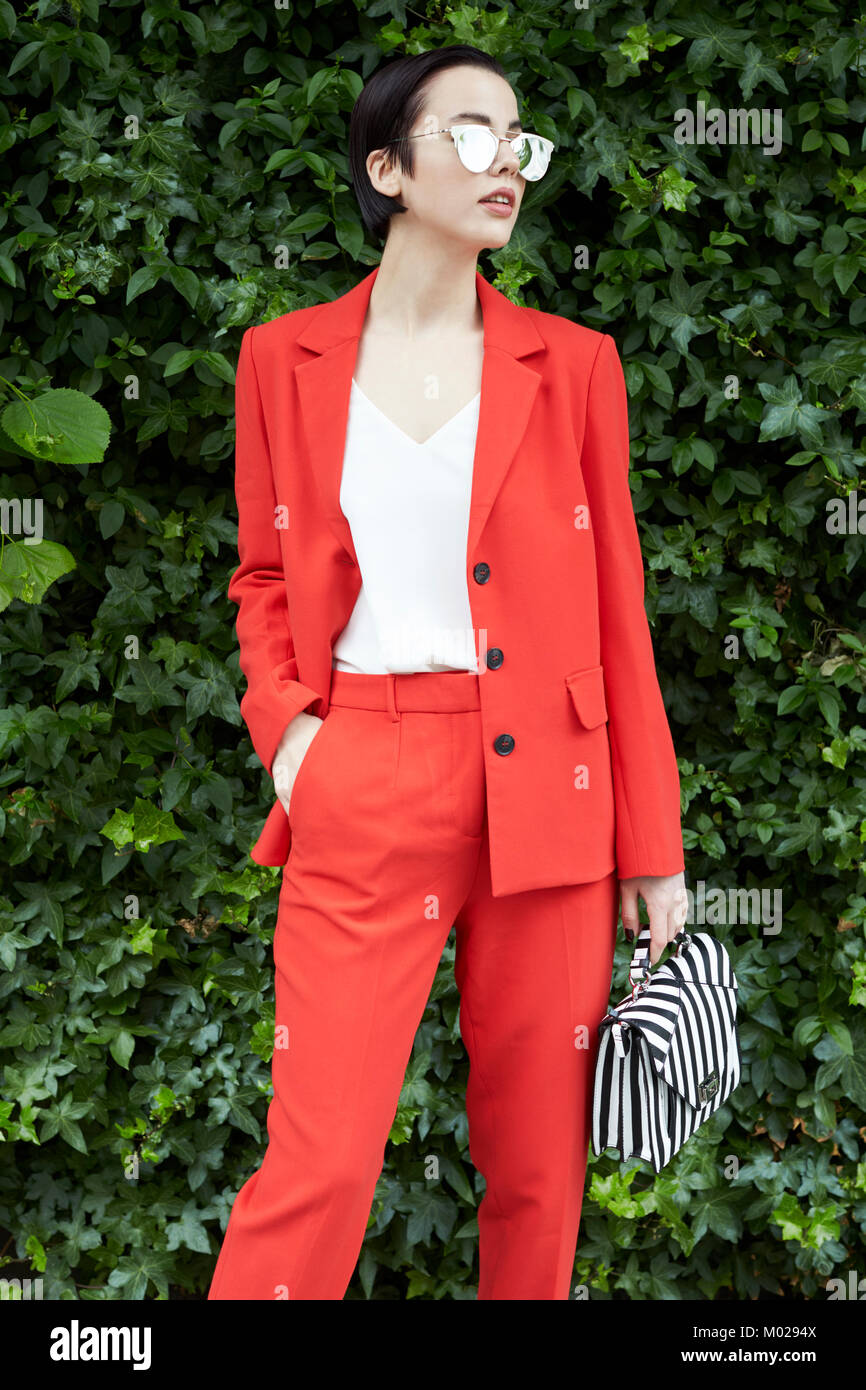 Chic woman in red suit in front of foliage looking away Stock Photo