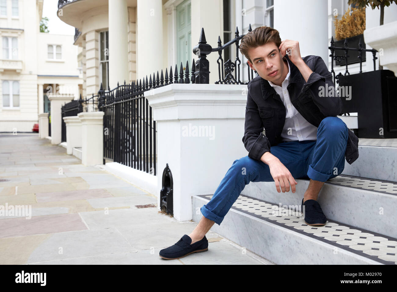 Young man sitting on steps outside house looking down street Stock Photo