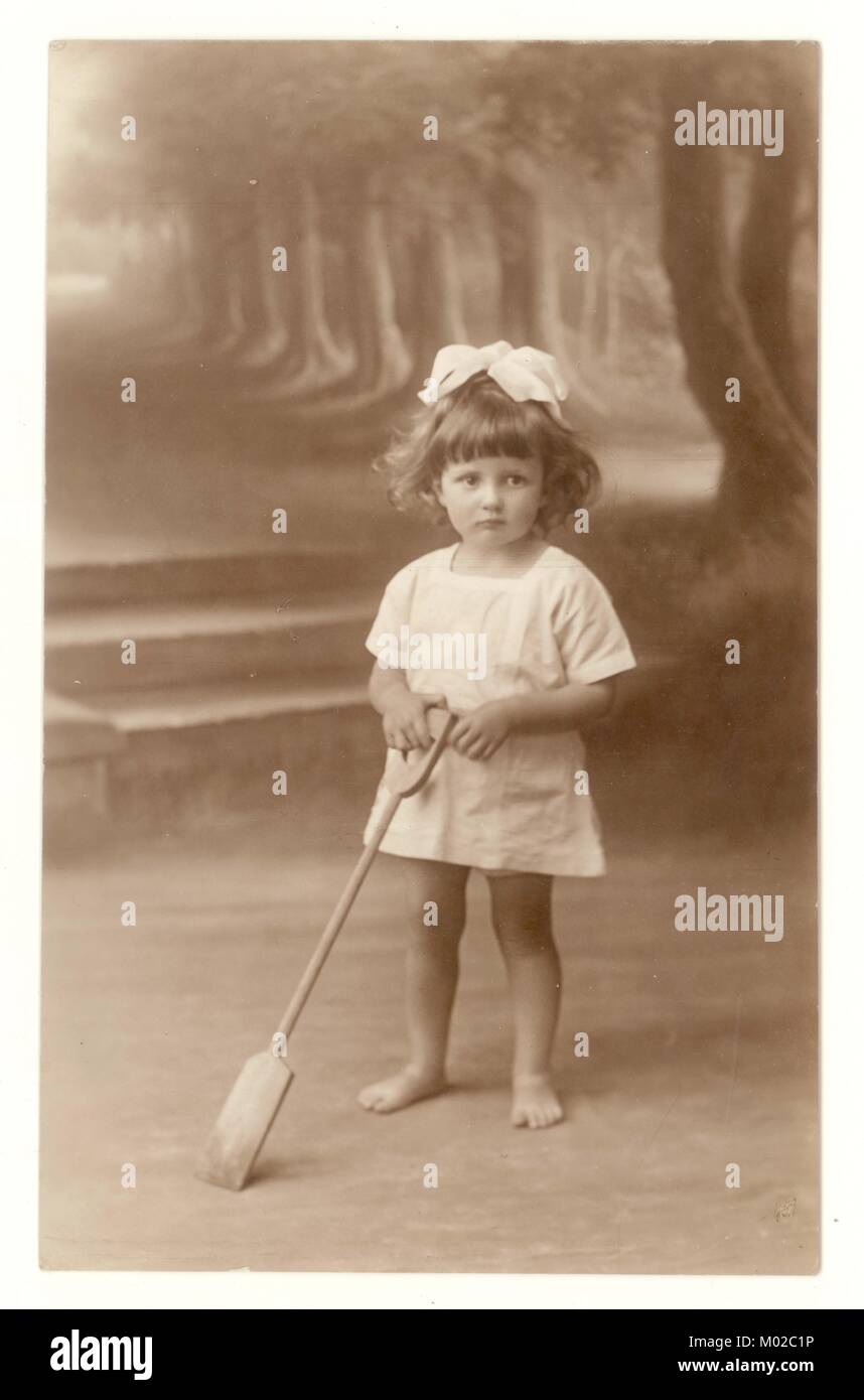 Original early 1900s studio portrait postcard with rural scene backdrop, of cute young girl with a ribbon in her hair, holding a spade, holiday photo, circa early 1920s, Southampton, Hampshire, England, UK Stock Photo