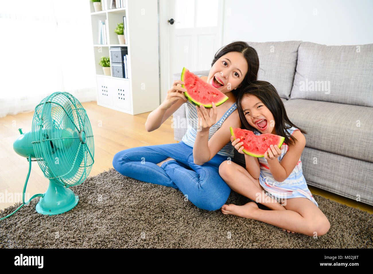 beautiful young lady with little girl eating watermelon looking at camera and sitting on living room floor enjoying cooling wind from electric fan. Stock Photo