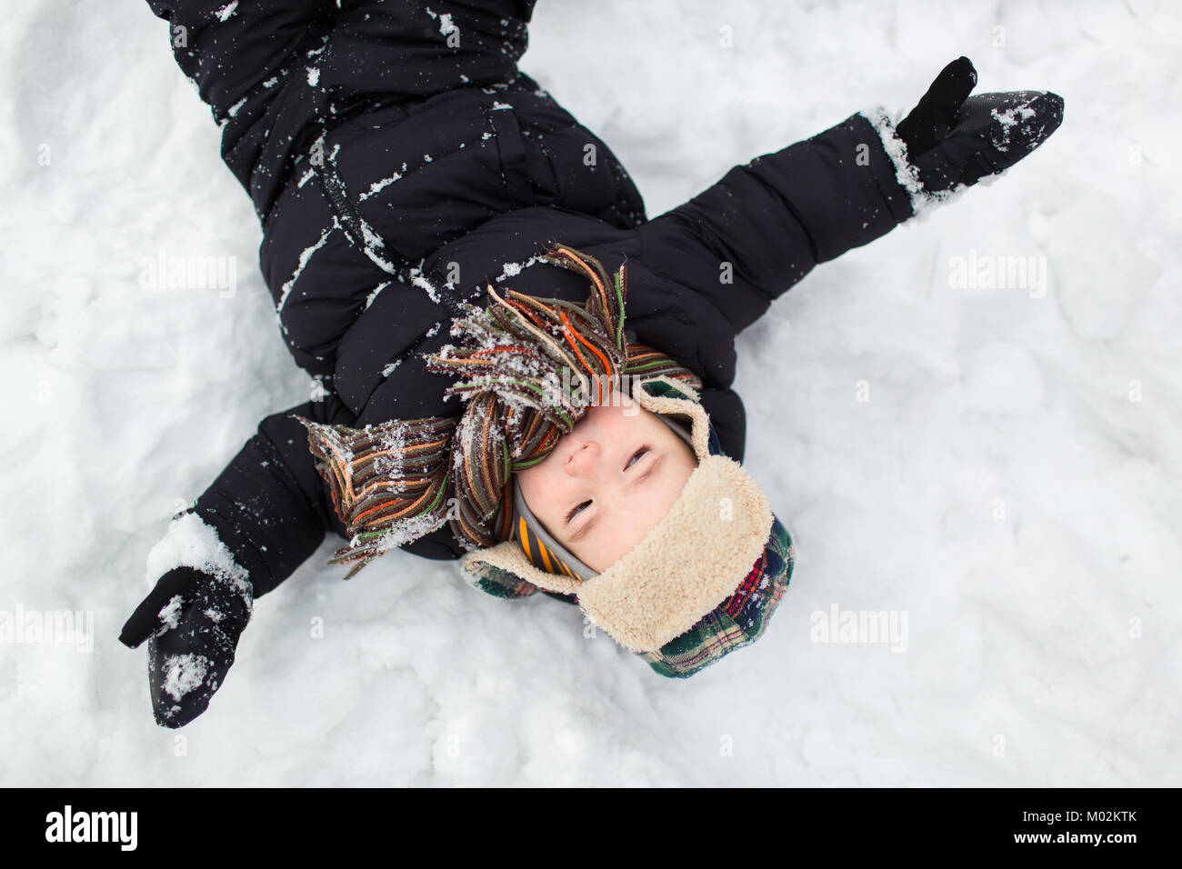 Top view of a little child dressed in warm clothes with his arms stretched wide in the snow. Boy making a snow angel. Stock Photo