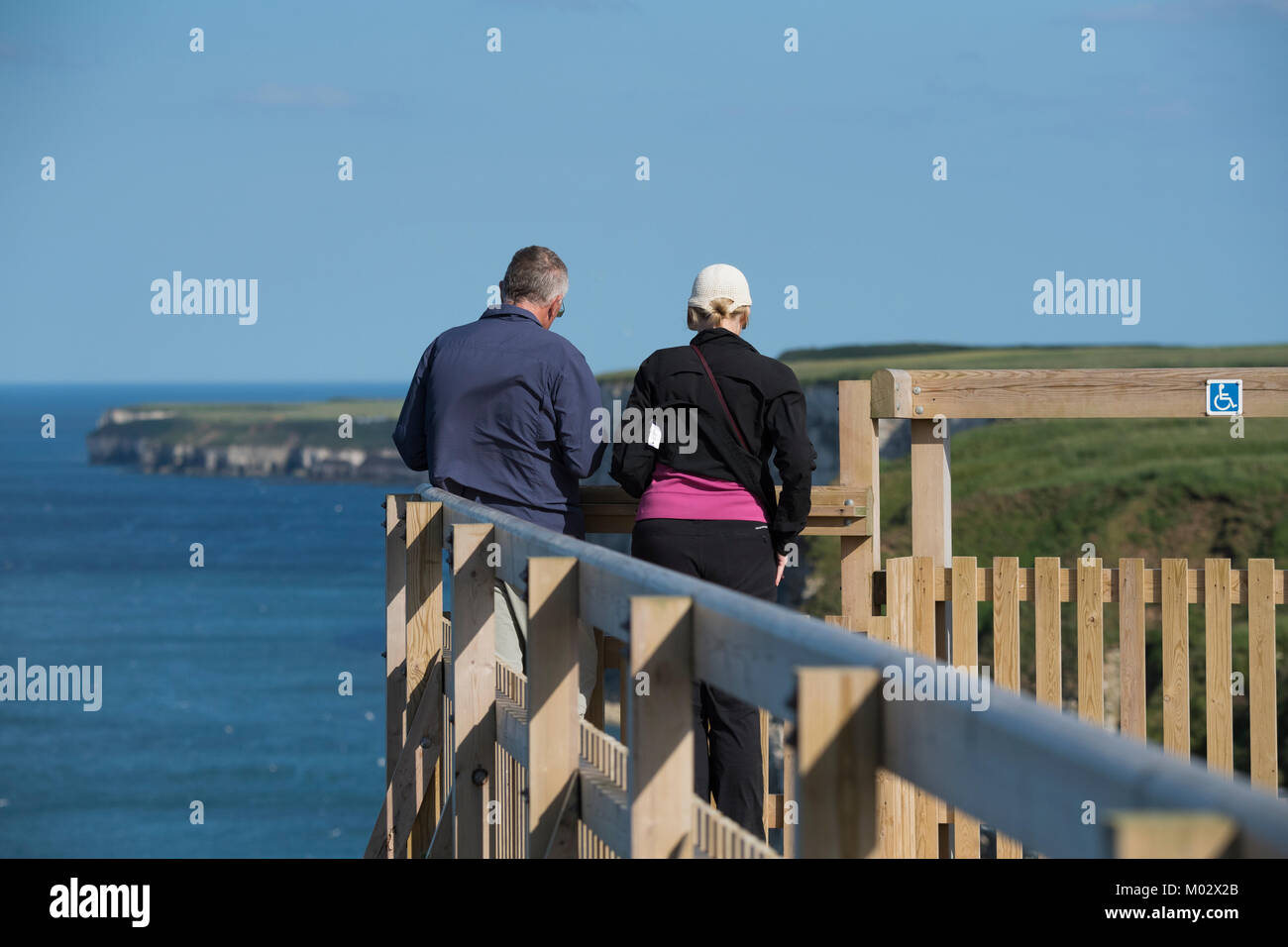 Cliff-top view for couple of people (birdwatchers) standing on viewing platform on sunny day - Bempton Cliffs RSPB reserve, East Yorkshire, England. Stock Photo
