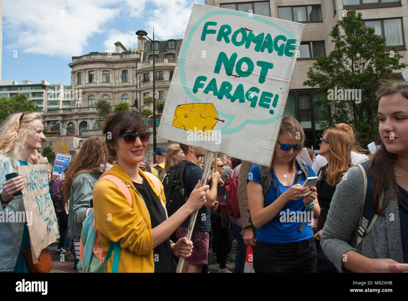 Demonstration against Brexit. Young woman, smiling, holds a placard 'Fromage not Farage' with illustration of cheese, with young protesters surroundin Stock Photo