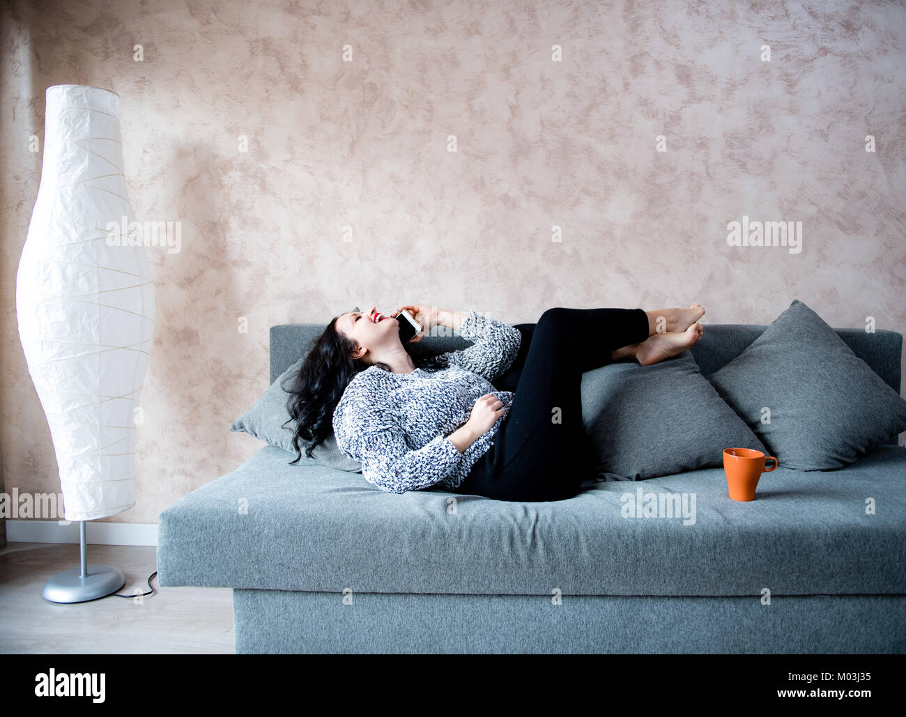 Young adult brunette woman laugh talking and using smart phone lying on couch at home Stock Photo