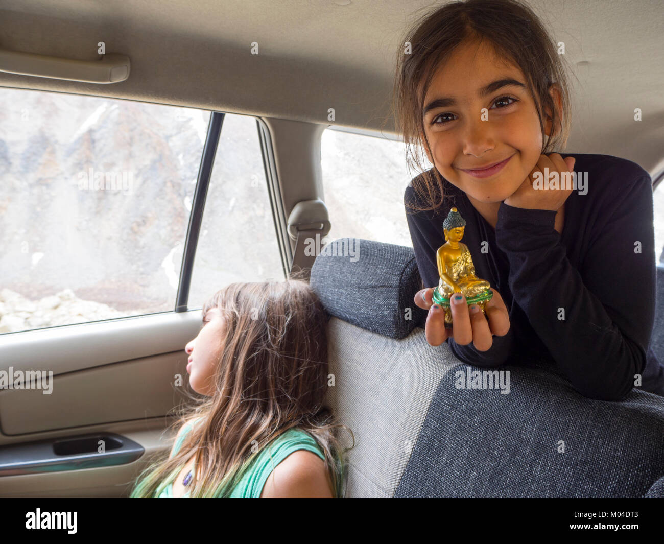 A beautiful girl holding a small statuette of Buddha while traveling by car on a trip to India. Her sister looks at the mountain view from the car win Stock Photo
