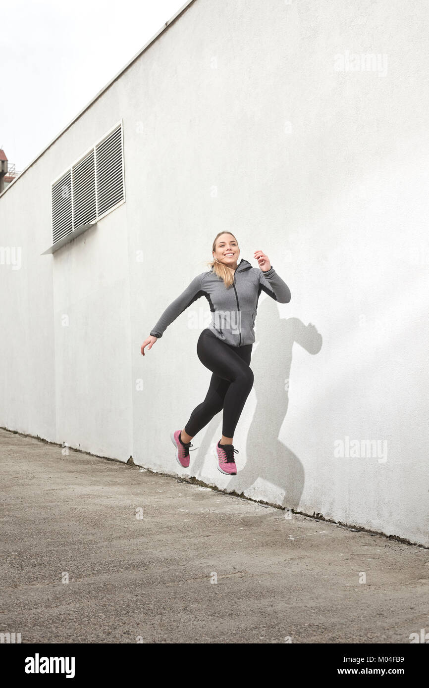 One young woman smiling happy, jumping in mid-air, outdoors, white wall behind, simple minimalistic, sport clothes. Stock Photo