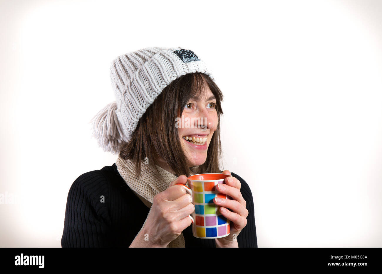 Inside portrait of happy female (side-on view) in woolly bobble ski hat, holding colourful mug of hot tea or coffee. Nice big natural smile. Stock Photo