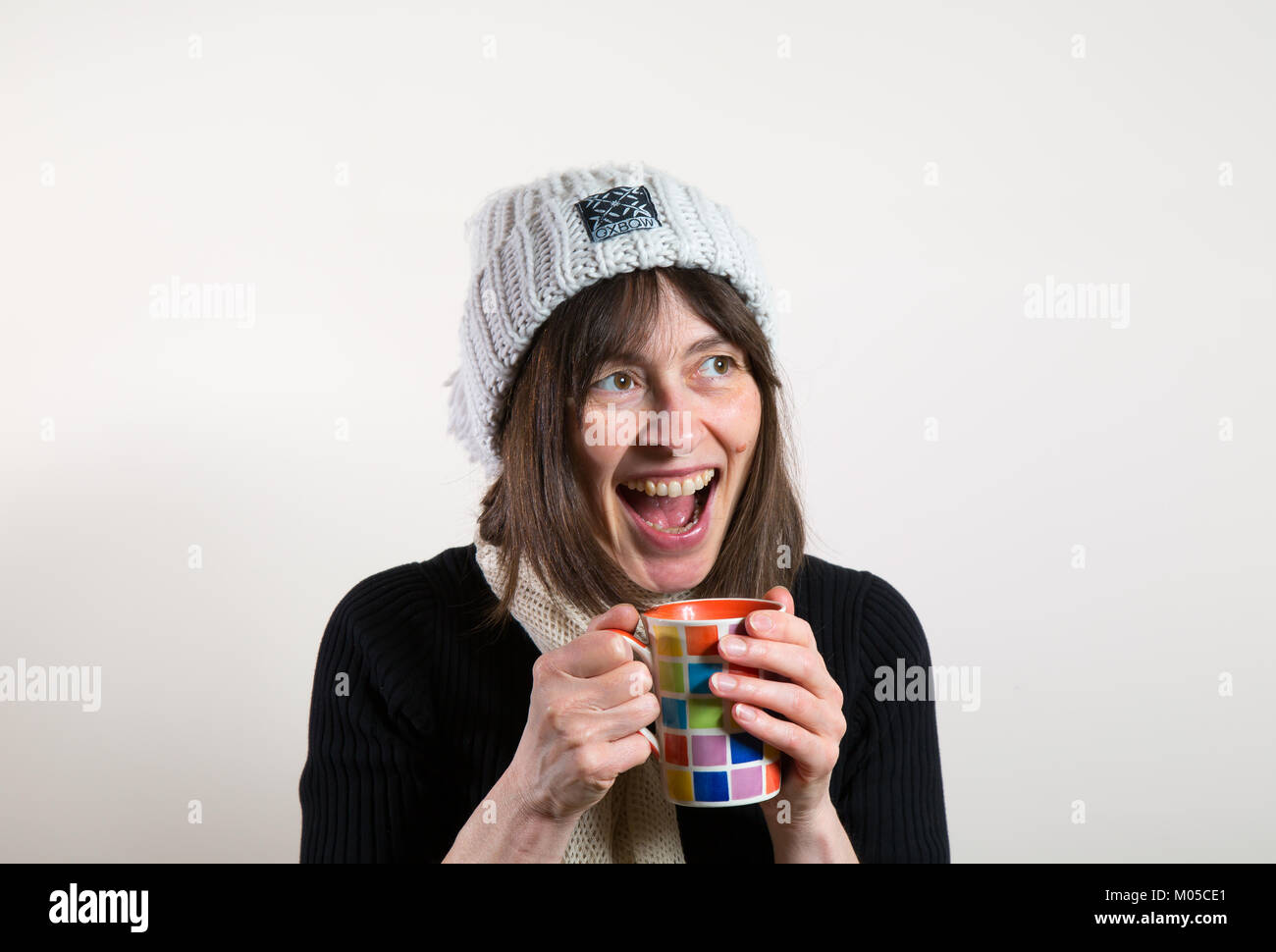 Portrait of happy female in woolly hat laughing (mouth open wide) holding mug, warming hands. Excited, cheesy grin showing teeth. Stock Photo