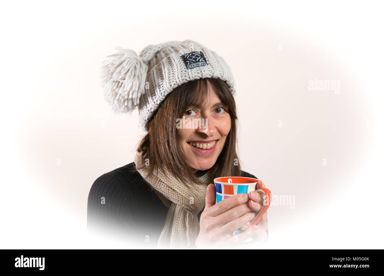 Indoor portrait of British woman looking at camera smiling in woolly hat & scarf, holding coffee and warming cold hands on hot mug. Big smile. Stock Photo