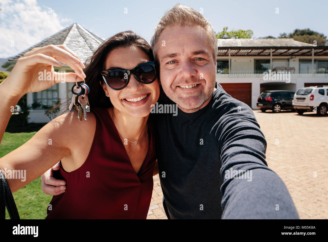 Happy couple with keys standing outside new home and taking a selfie ...