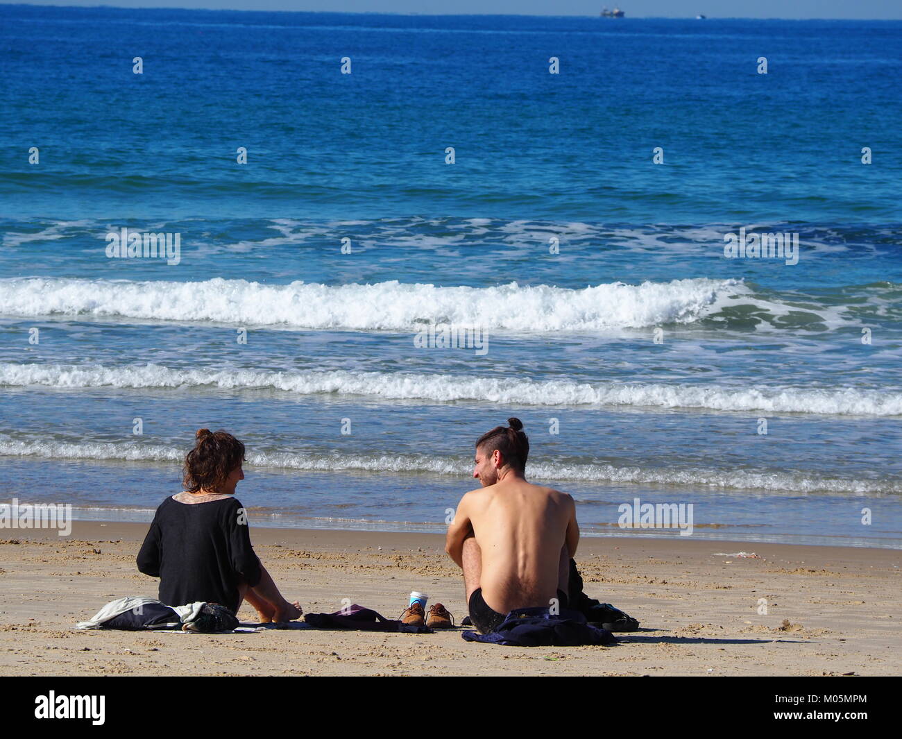 two Friends sitting on the beach sand on a perfect clear day and having a conversation with each other Stock Photo