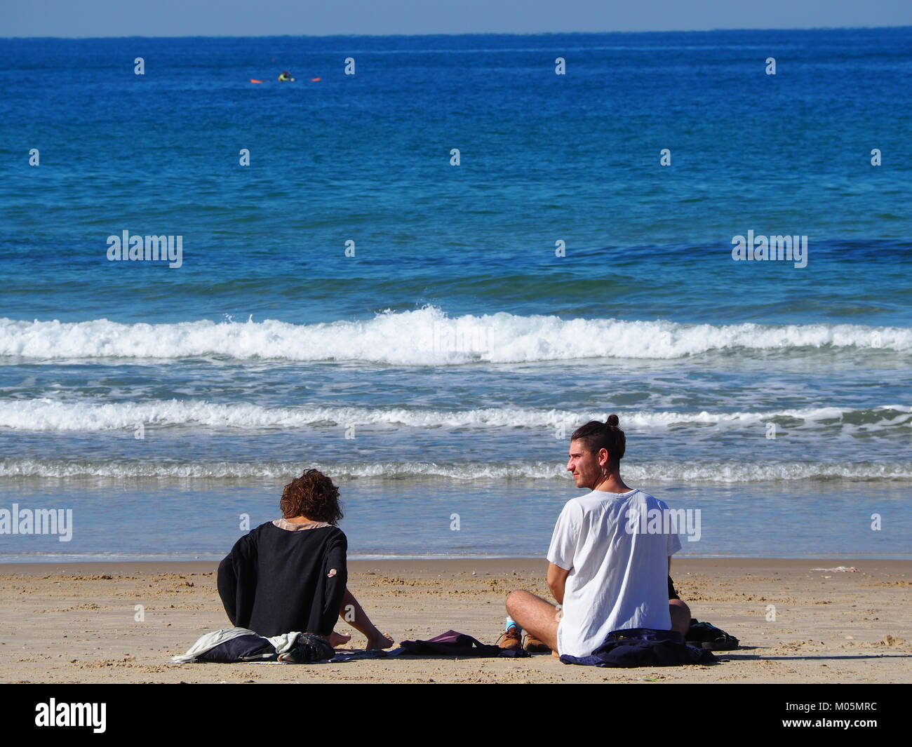 two Friends sitting on the beach sand on a perfect clear day and having a conversation with each other Stock Photo