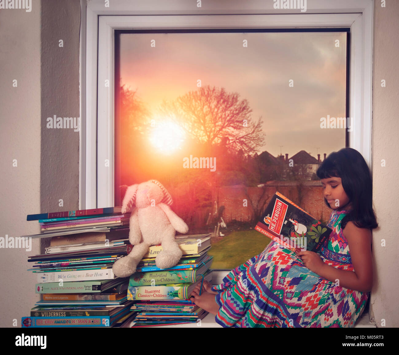 Little girl in colourful dress reading a book by the window against a sunset in the background Stock Photo