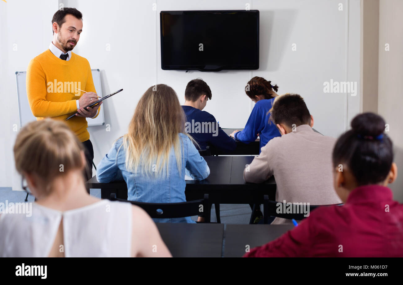 Positive friendly smiling teacher monitoring students’ work during examination test in class Stock Photo