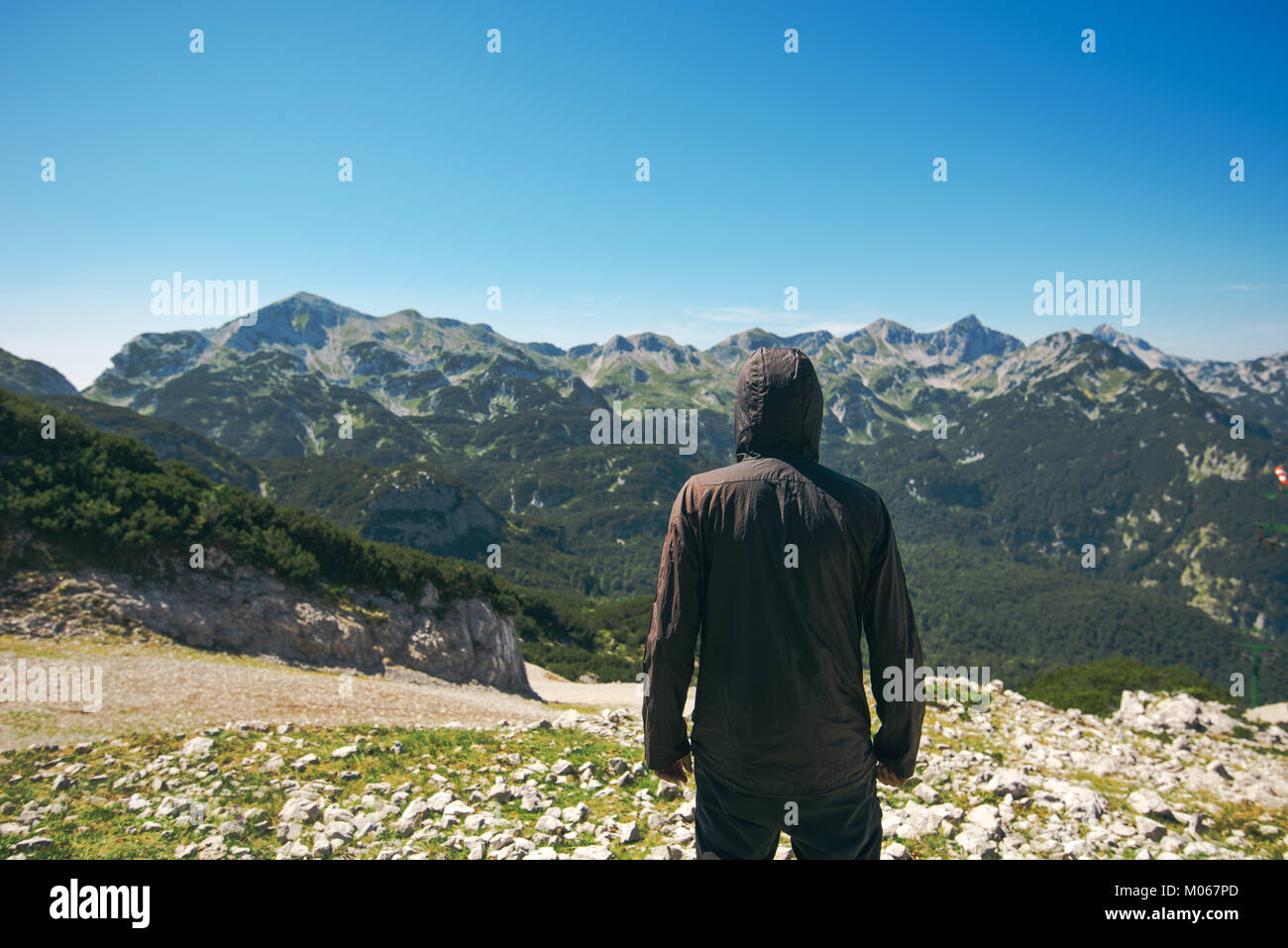 Mountain hiker at high viewpoint looking at the valley. Male tourist person in hooded jacket at mountain top enjoying the view. Stock Photo