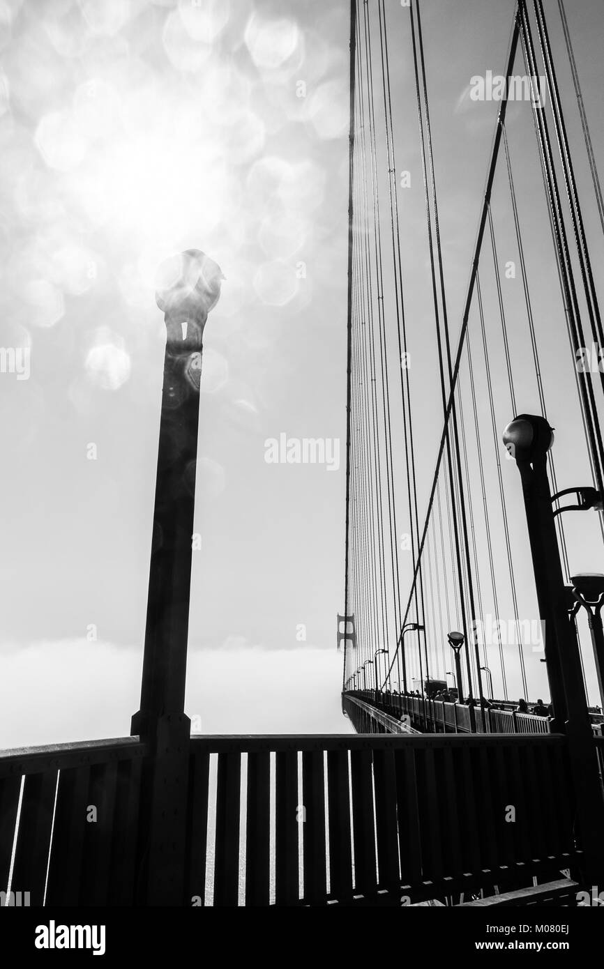 Golden Gate Bridge rising over dense fog below, with sun flare bouncing off a lamp post on the bridge. Close up view. Black and white monochrome. Stock Photo