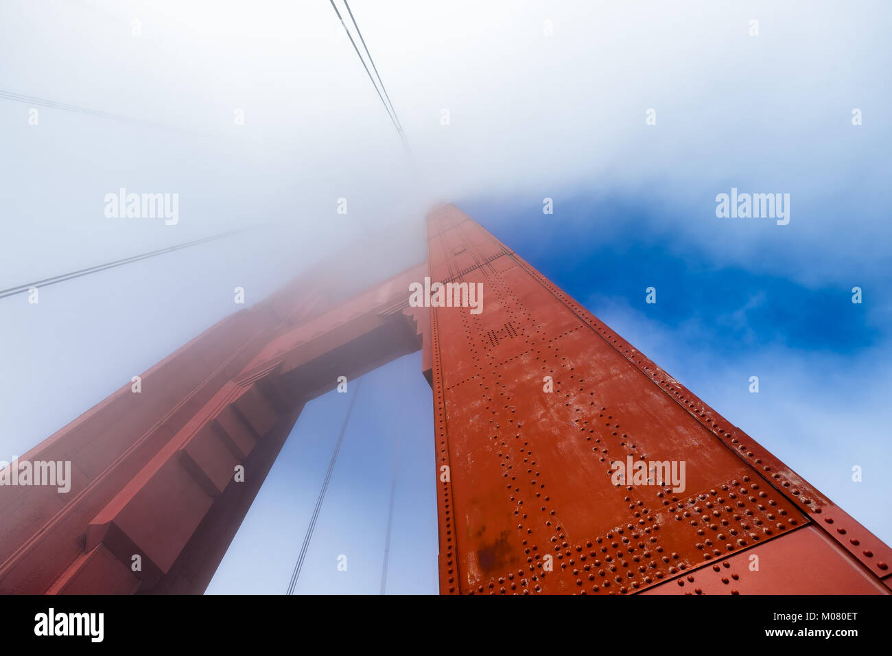 Golden Gate Bridge tower looking up from the base. Close up of rivets in the orange metal. Top of tower disappears into fog. Patches of blue sky. Stock Photo