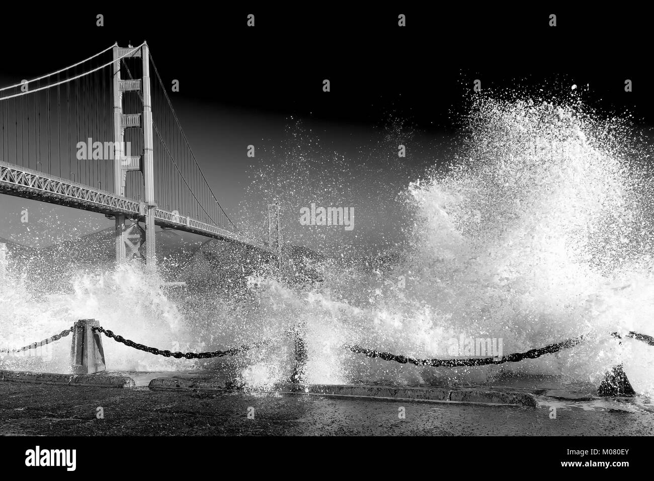 Golden Gate Bridge. Huge waves crash over the sea wall below the bridge in the foreground . Black and white monochrome with dark sky. Copy Space. Stock Photo