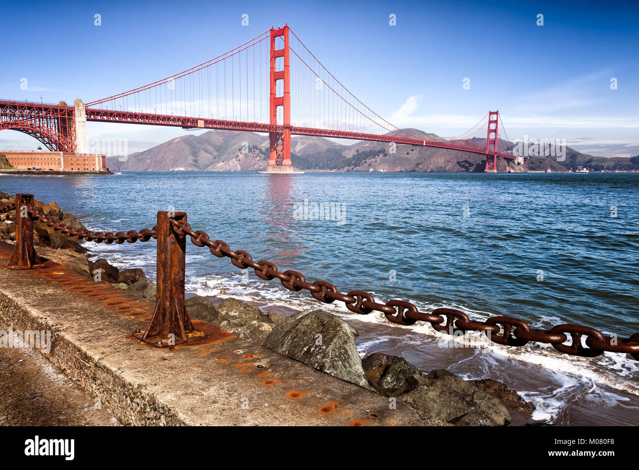Golden Gate Bridge view. Foreground at water's edge, with a heavy rusty chain guardrail that echoes the shape of the bridge. Vintage color. Stock Photo