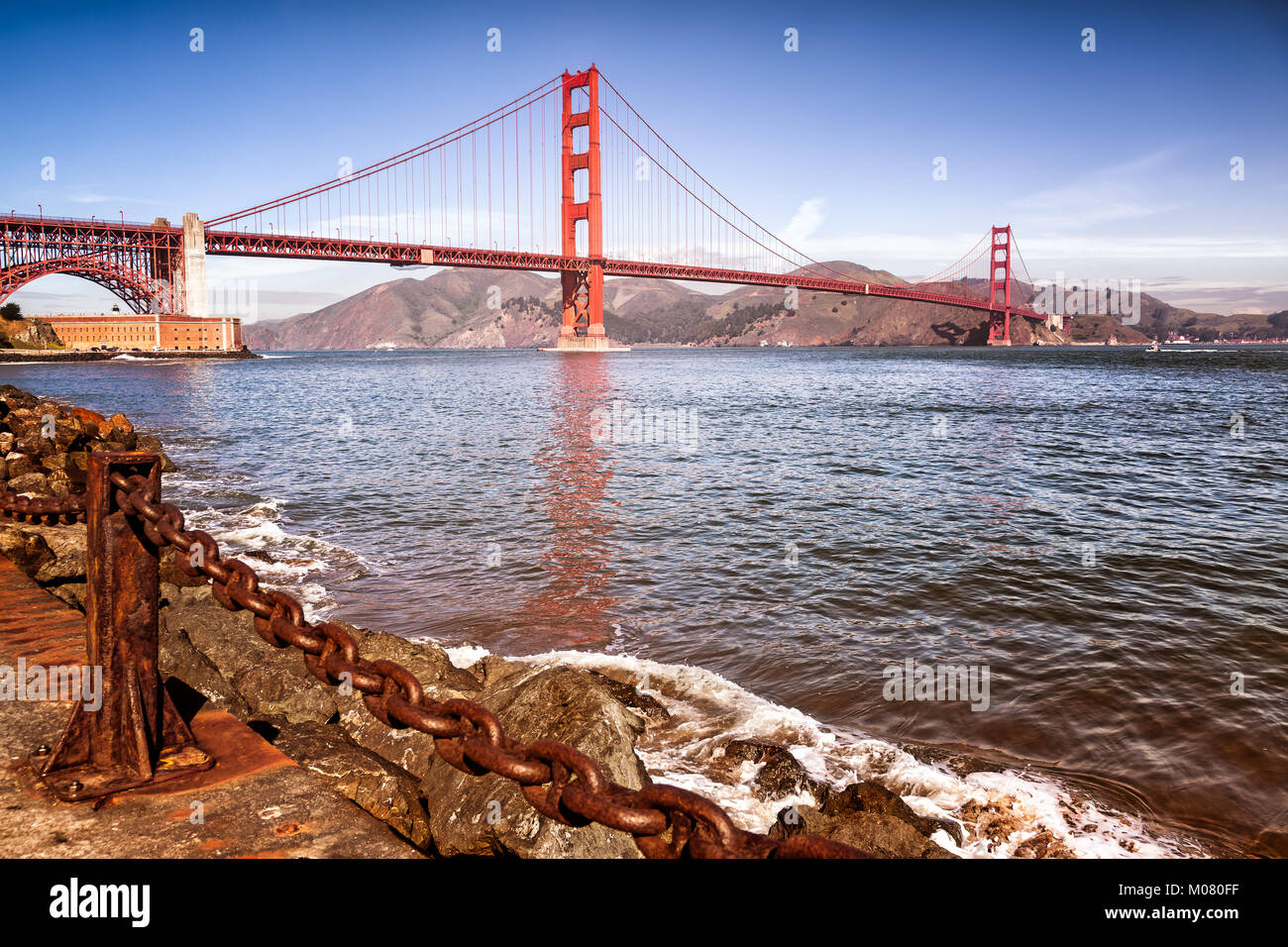 Golden Gate Bridge. Foreground of water's edge, rocky boulders and a heavy rusty chain guardrail. Stock Photo