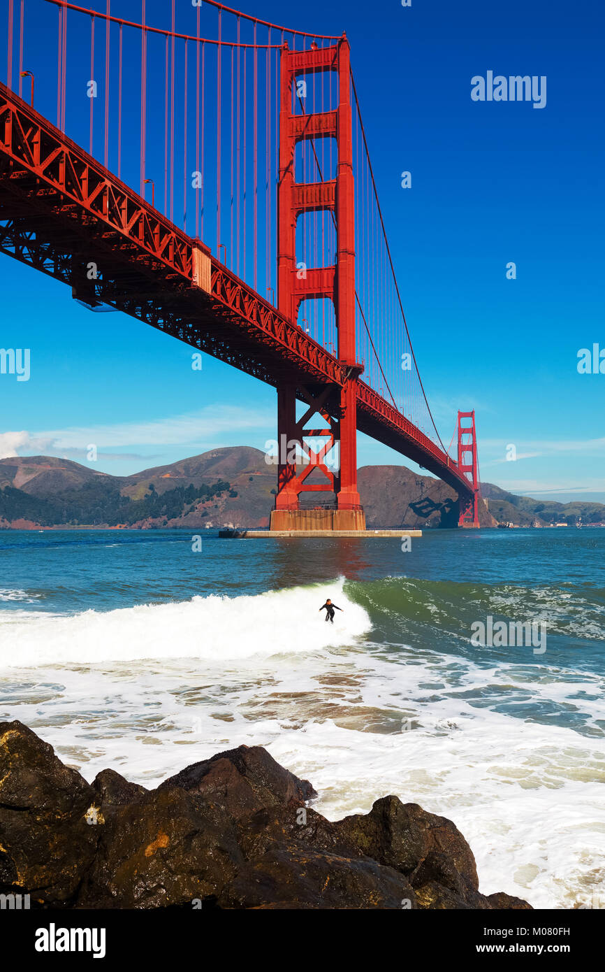 Golden Gate Bridge surfer rides a wave under the span toward the rocky boulders at the base of the bridge Stock Photo