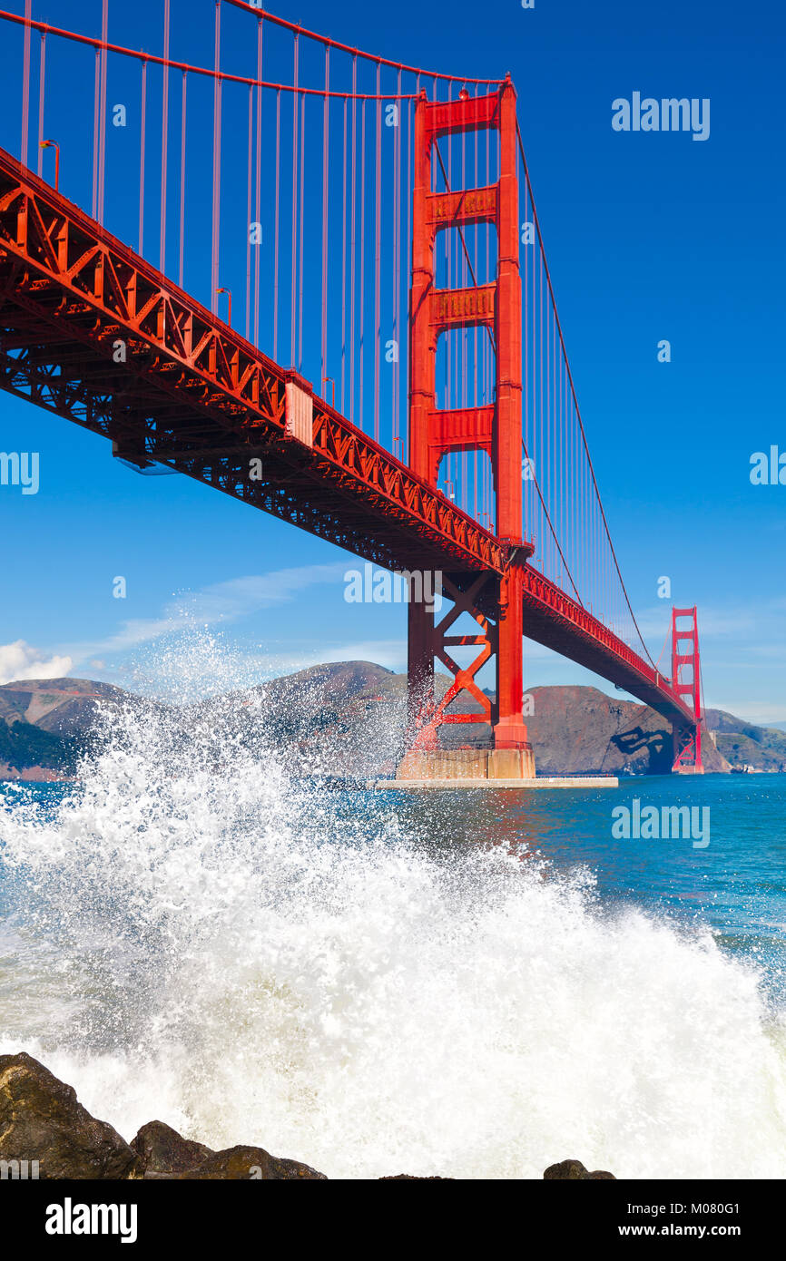 Golden Gate Bridge viewed from below with a big ocean wave spray in the foreground. Bright blue sky and colorful bridge. Vertical. Stock Photo