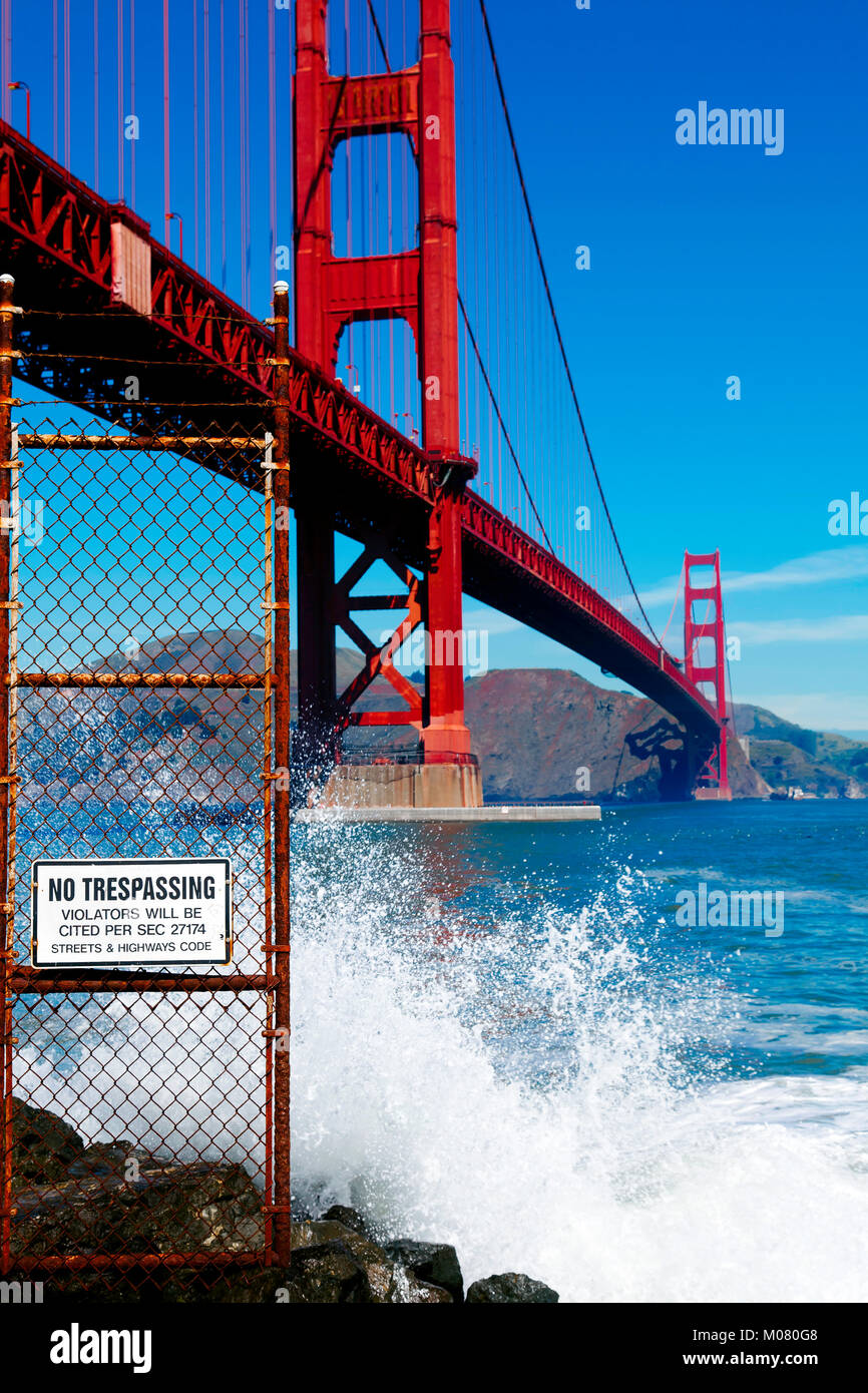 Golden Gate Bridge seen from below. Sign that reads No Trespassing  next to dangerous slippery rocks and surf. Stock Photo