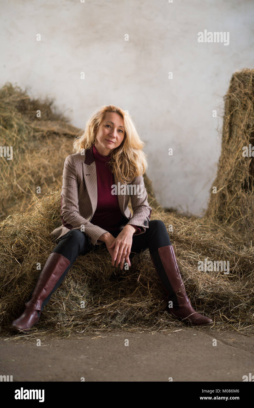 Blonde woman in a jacket and boots is sitting on the hay Stock Photo