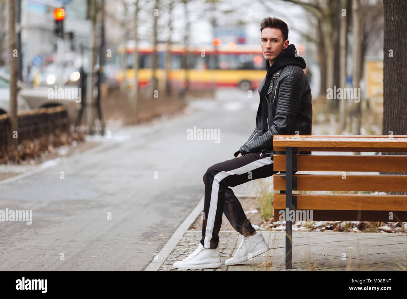 Modern outfit. Fashionable man sit on the bench and look around. Autumn and winter clothing style. Jacket with white sneakers. Stock Photo