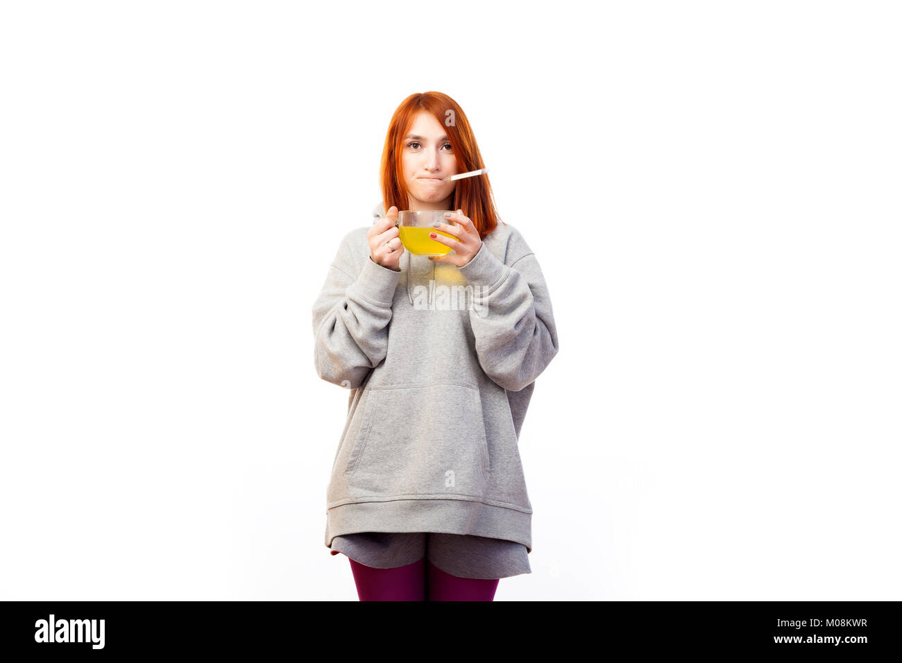 A young red-haired woman in a gray sweatshirt is sad because she has the flu, measures the temperature with a thermometer and drinks from a large tran Stock Photo