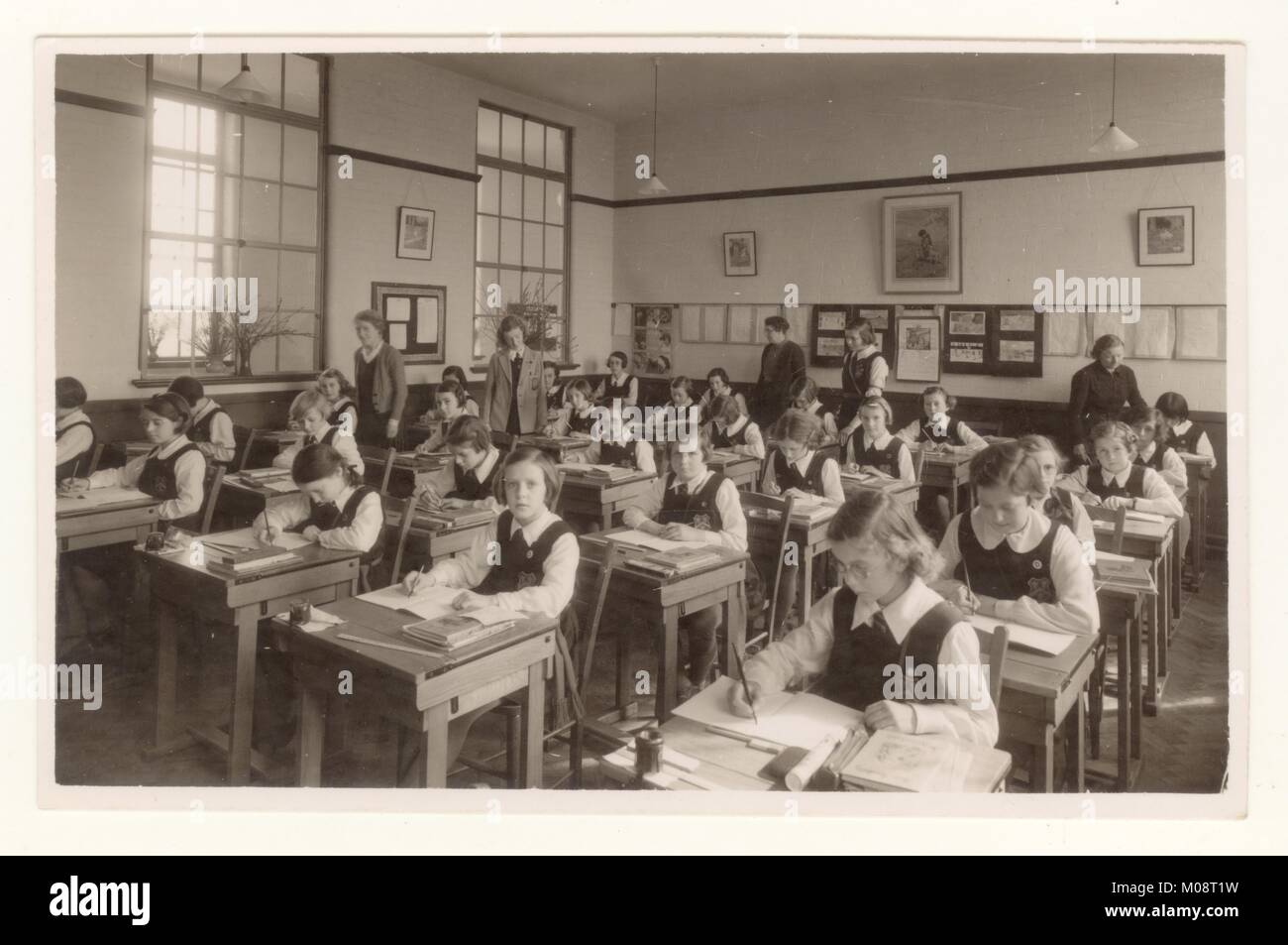 Postcard photograph of school classroom of girls writing at desks using pen and ink from an ink pot, with teachers and class monitors helping, circa 1930s, U.K. Stock Photo