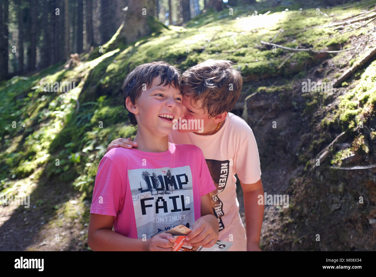 Two boys, brothers, walking and laughing as one of them tells a secret with his hand on a shoulder of the other in a forest. Stock Photo