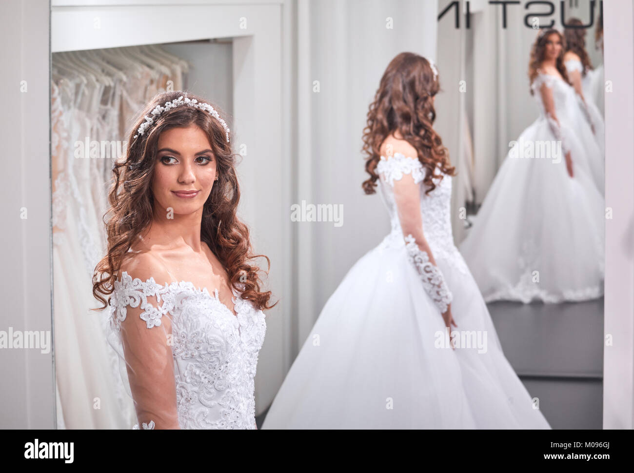 one young bride, looking at self, wearing gown, in mirror image back, front view, bridal salon. Stock Photo