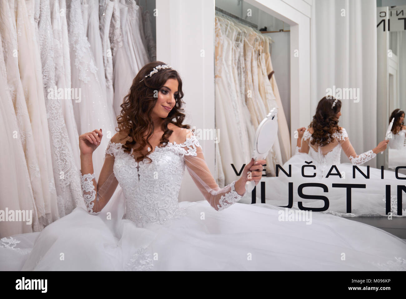 one person, young bride, looking at herself in mirror, bridal salon, wearing gown. Stock Photo