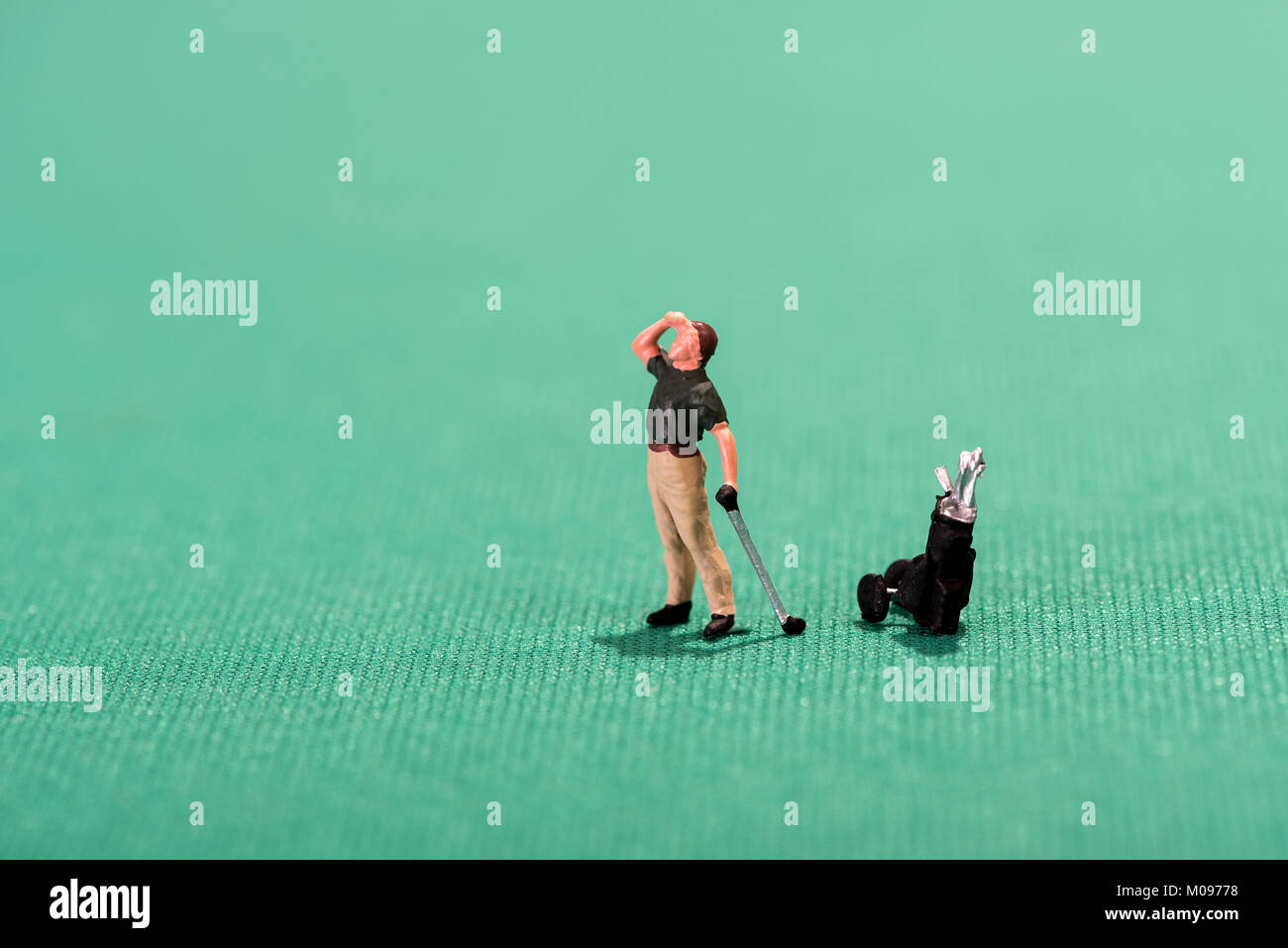 Miniature figure of a golfer peering into the distance after playing a stroke to see where his ball lands with his bag of clubs on a green background Stock Photo