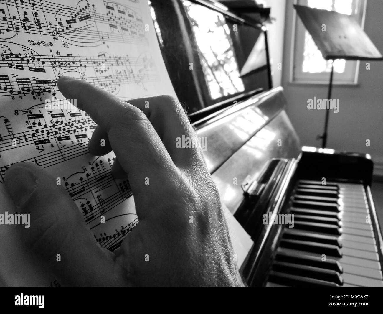 A male hand is on a piano sheet music touching the paper like he is playing. An acoustic piano and a music stand in the background. Stock Photo