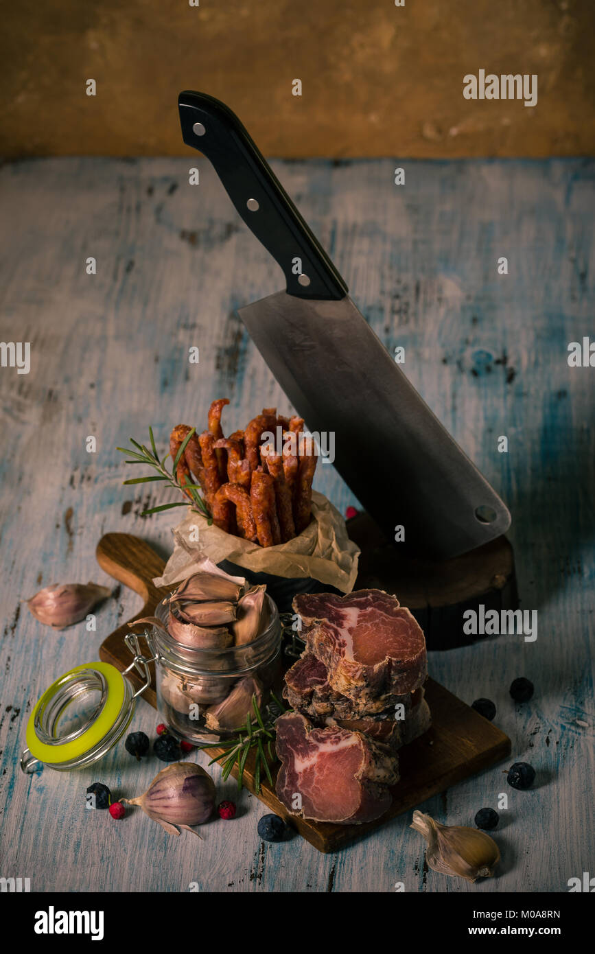 Vertical photo of stacked pieces of dried smoked meat from italian wild boar. Meat is on vintage chopping board and blue wooden table with sausages, g Stock Photo