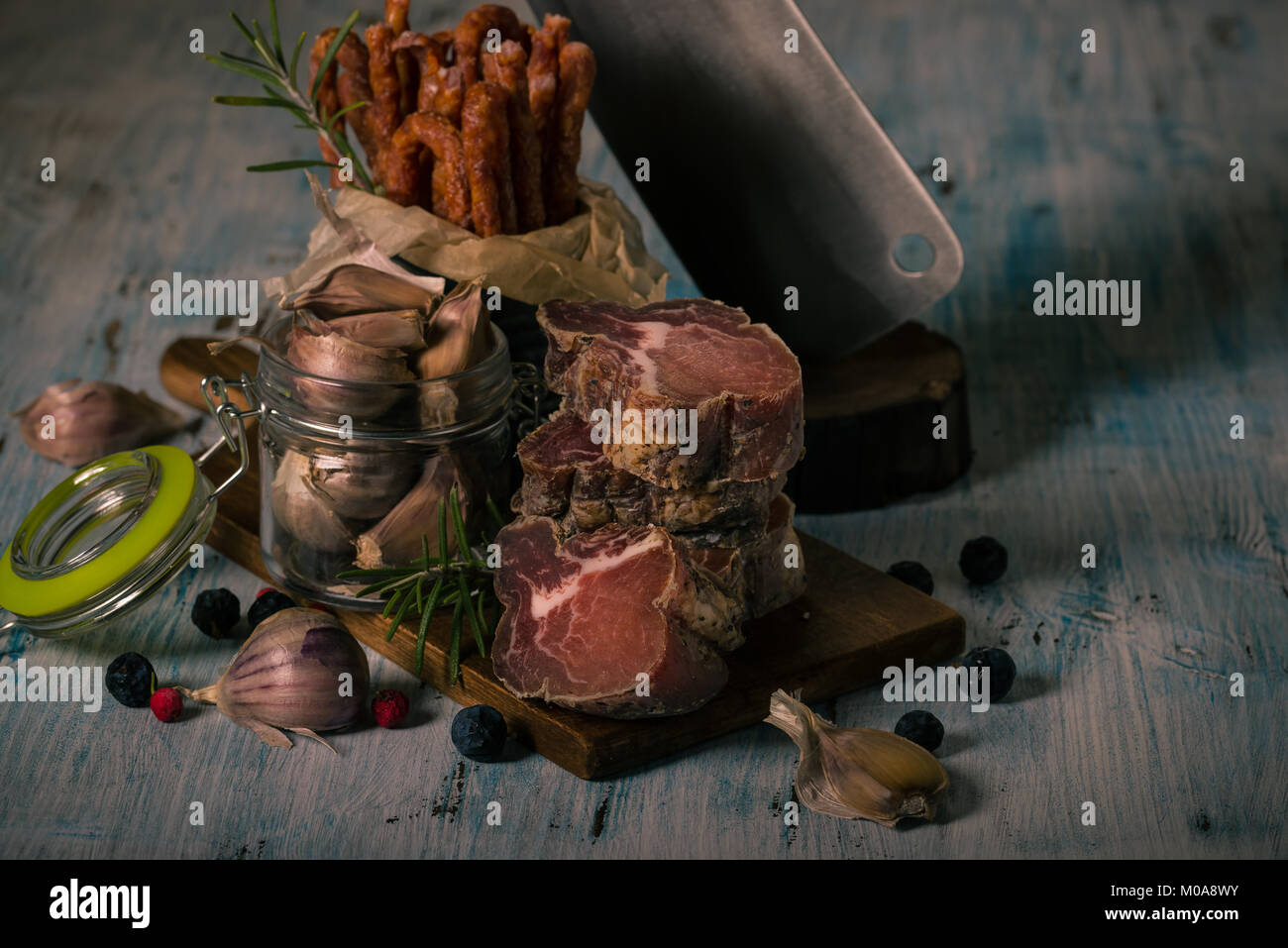 Horizontal photo of stacked pieces of dried smoked meat from italian wild boar. Meat is on vintage chopping board and blue wooden table with sausages, Stock Photo