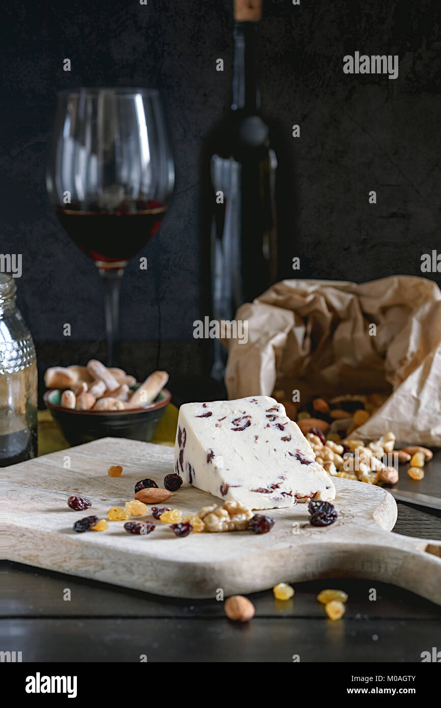 Wensleydale cheese with cranberries, red wine, honey, nuts, raisins on wooden cutting board. Black concrete background. Selective focus. Stock Photo