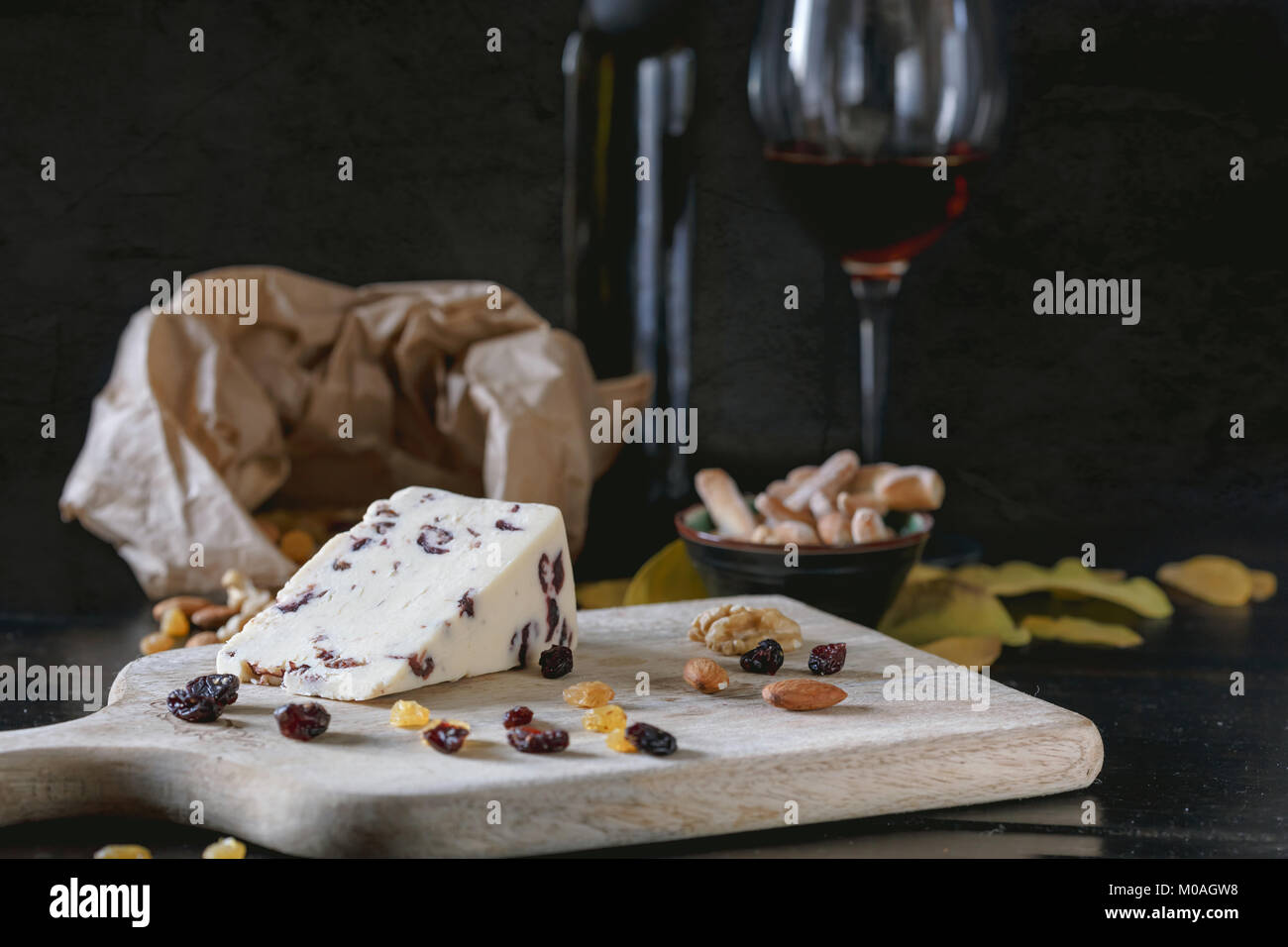 Wensleydale cheese with cranberries, red wine, honey, nuts, raisins on wooden cutting board. Black concrete background. Selective focus. Stock Photo