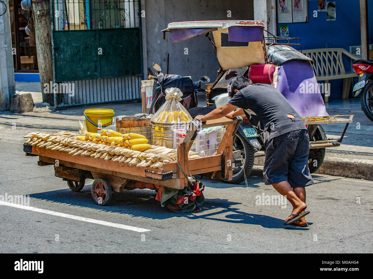 A Filipino street food vendor works hard pushing his heavy food cart full of corn up a road in Agoo, Luzon Island, Philippines. Stock Photo