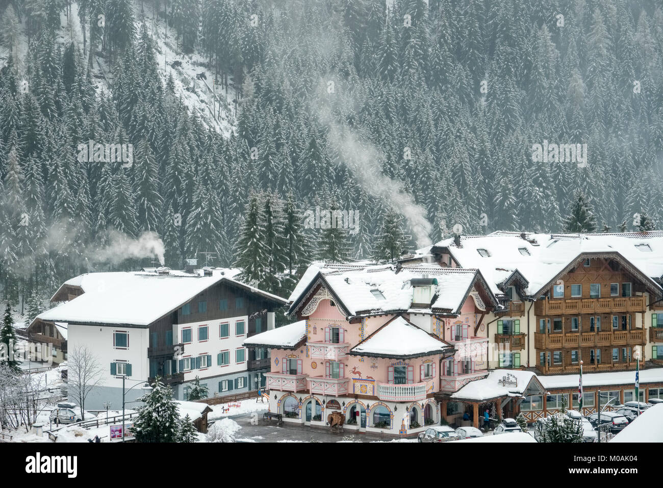 Pink, highly decorated hotel in Campitello with smoke from the chimneys and the tree-covered mountain with snow behind. Stock Photo