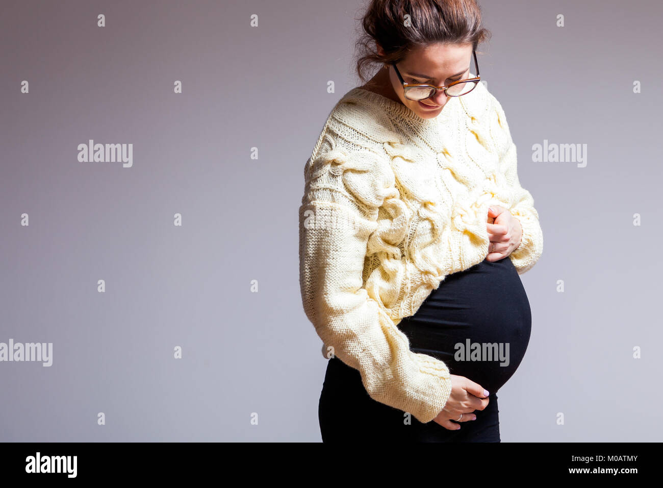 Young dark-haired beautiful woman in late pregnancy with glasses and knitted milk-color sweater posing and holding her stomach on a white isolated bac Stock Photo