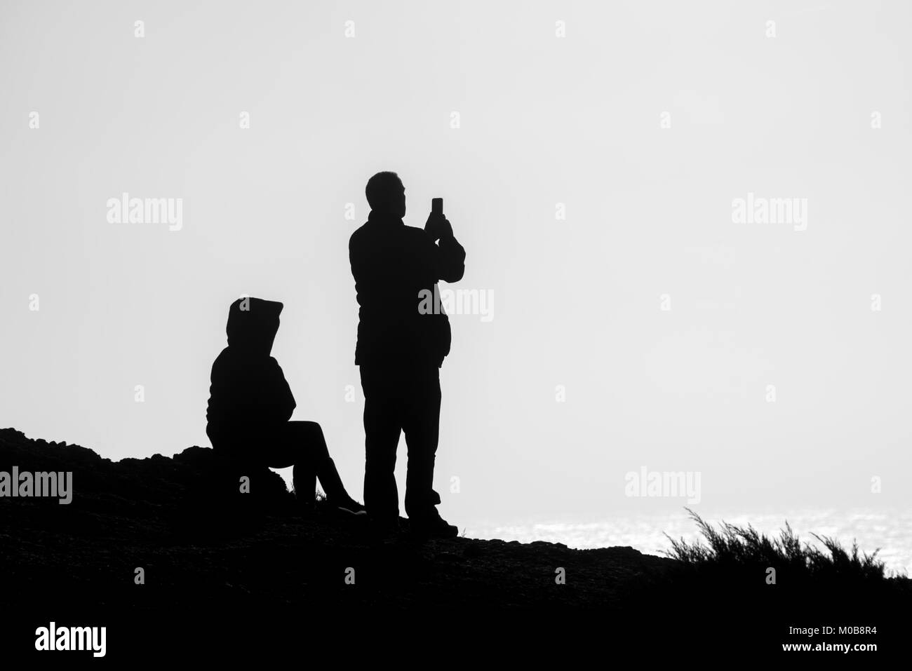 silhouette of a couple on top of a cliff taking a photo with a smart phone. Stock Photo
