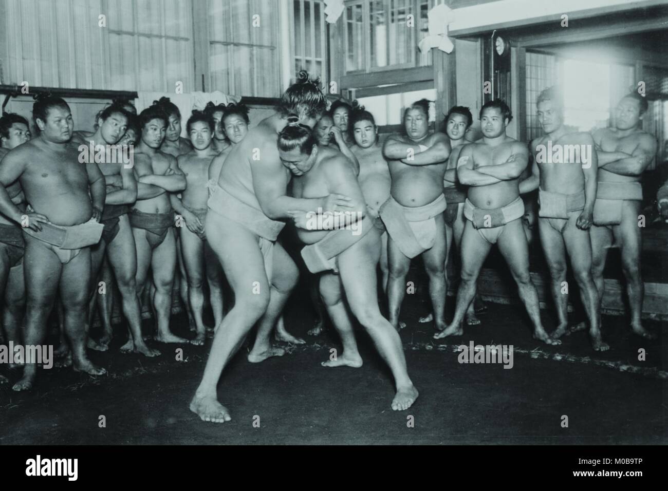 Two Japanese Sumo Wrestlers attempt to get the advantage over their opponent while the full  cadre of fellow sumo look on. Stock Photo