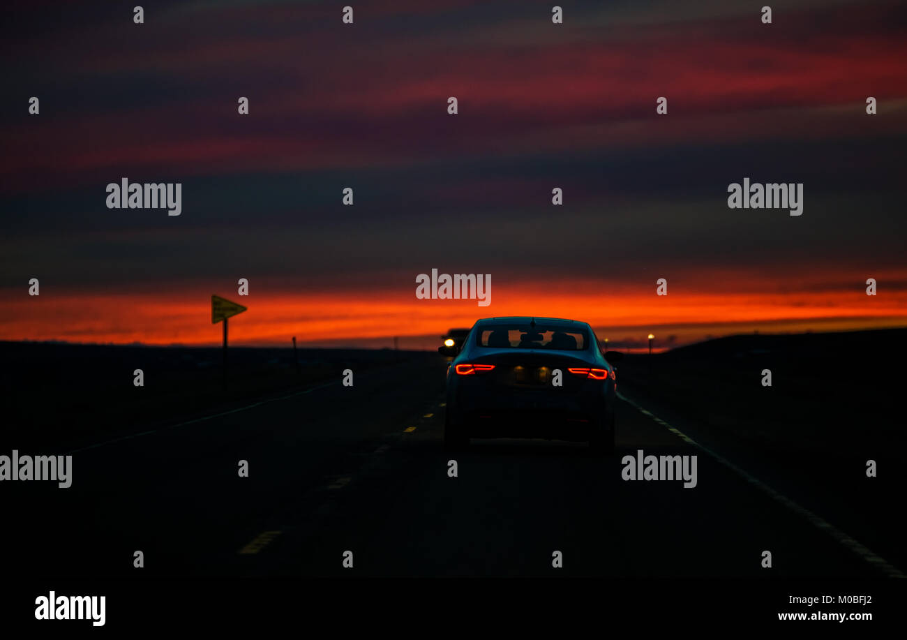 Red taillights of a travelling car on a highway against a colorful sunset in a rural landscape Stock Photo