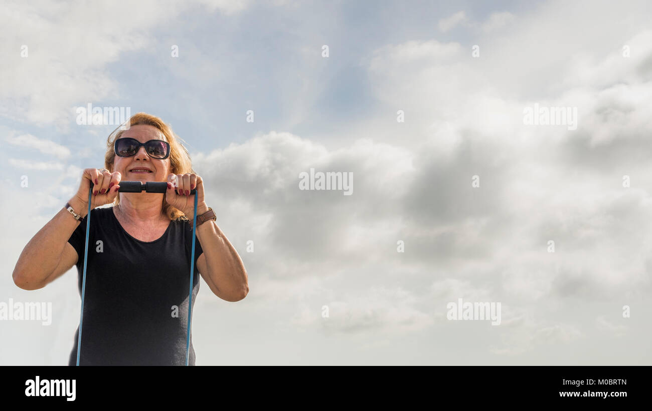 Model Released: Mature woman (70-75) exercising with a stretch cord against blue sky copy space Stock Photo