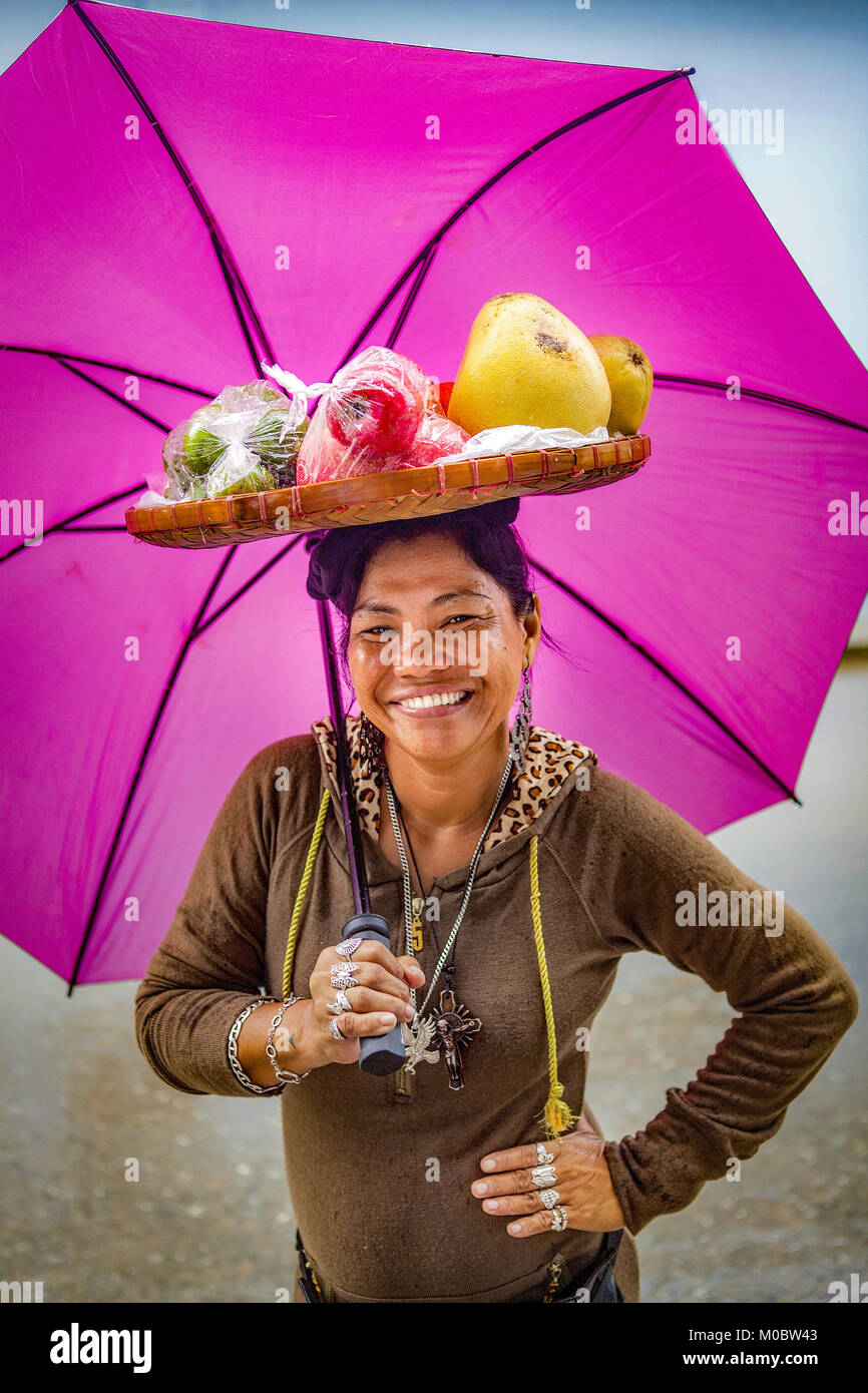 Filipino woman with a basket of fruit on her head and a bright, magenta colored umbrella smiles at the camera in Baloy Long Beach, Luzon, Philippines. Stock Photo