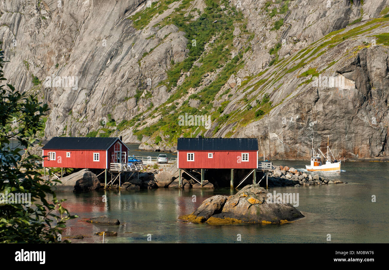 Typical rorbu cottages in Nusfjord and a fishing boat cruising out into the Norwegian sea. Stock Photo